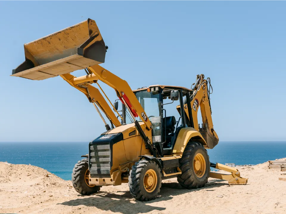 CAT 416 backhoe loader holding its front bucket high on a sandy ridge above the sea