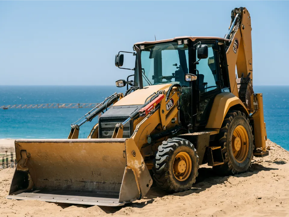 Yellow CAT 428 backhoe loader with its front bucket resting on sand and the ocean behind it