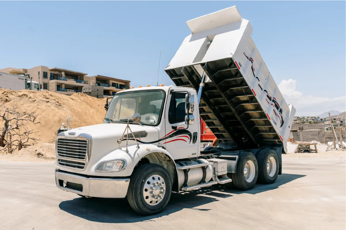 White Freightliner M2 112 seen from the front corner with its empty white dump body tipped high on the hoist, a mound of sand and a dry tree behind it
