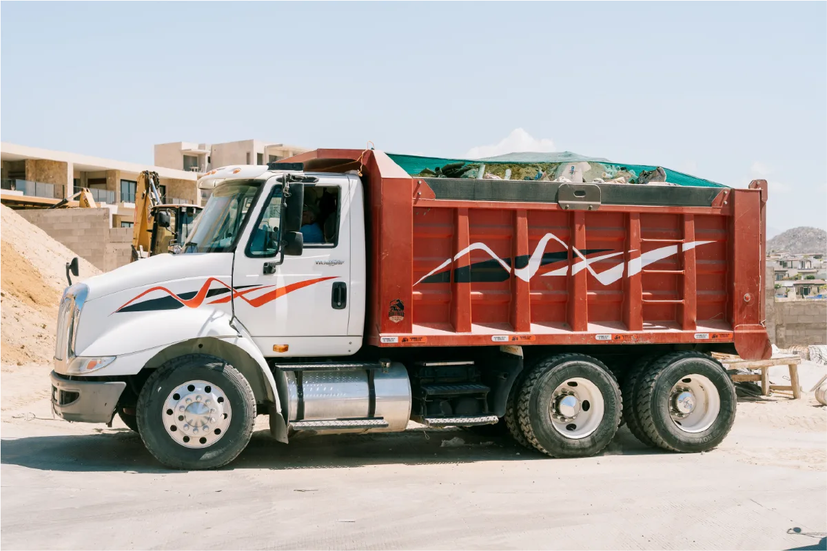 International TranStar dump truck parked beside a sand stockpile, its red ribbed body full and covered, a yellow backhoe and new villas in the background
