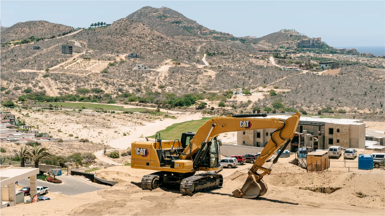 CAT 320GC excavator working a hillside plot that overlooks a green valley, with low buildings, parked cars and desert mountains in the distance