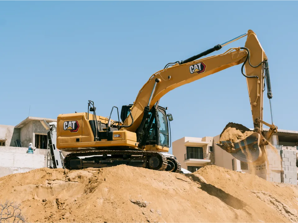 CAT 320GC excavator emptying a bucket of sand onto a mound beside unfinished concrete buildings