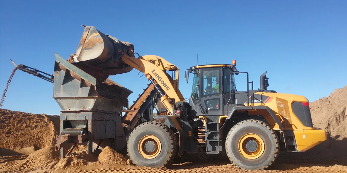 LiuGong 856H loader seen side on with its arms fully raised, tipping a bucket of sandy material into a steel hopper that feeds a conveyor, an operator in a high visibility vest at the controls