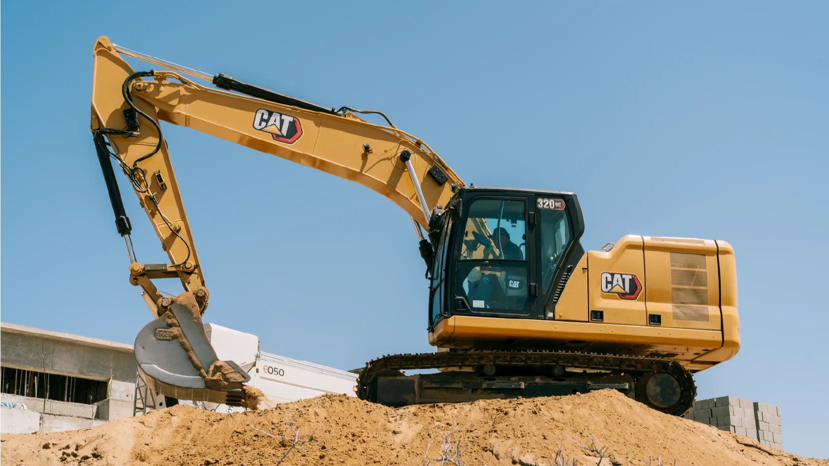 CAT 320GC crawler excavator with an operator in the cab, standing on a mound of sand at a building site under a clear sky