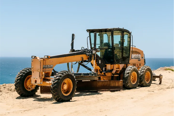 The CASE 865B grader seen from its front quarter on a sandy clifftop, showing the 865B lettering on the front frame, the glass cab, the blade under the machine and the ripper teeth at the rear, with open sea behind