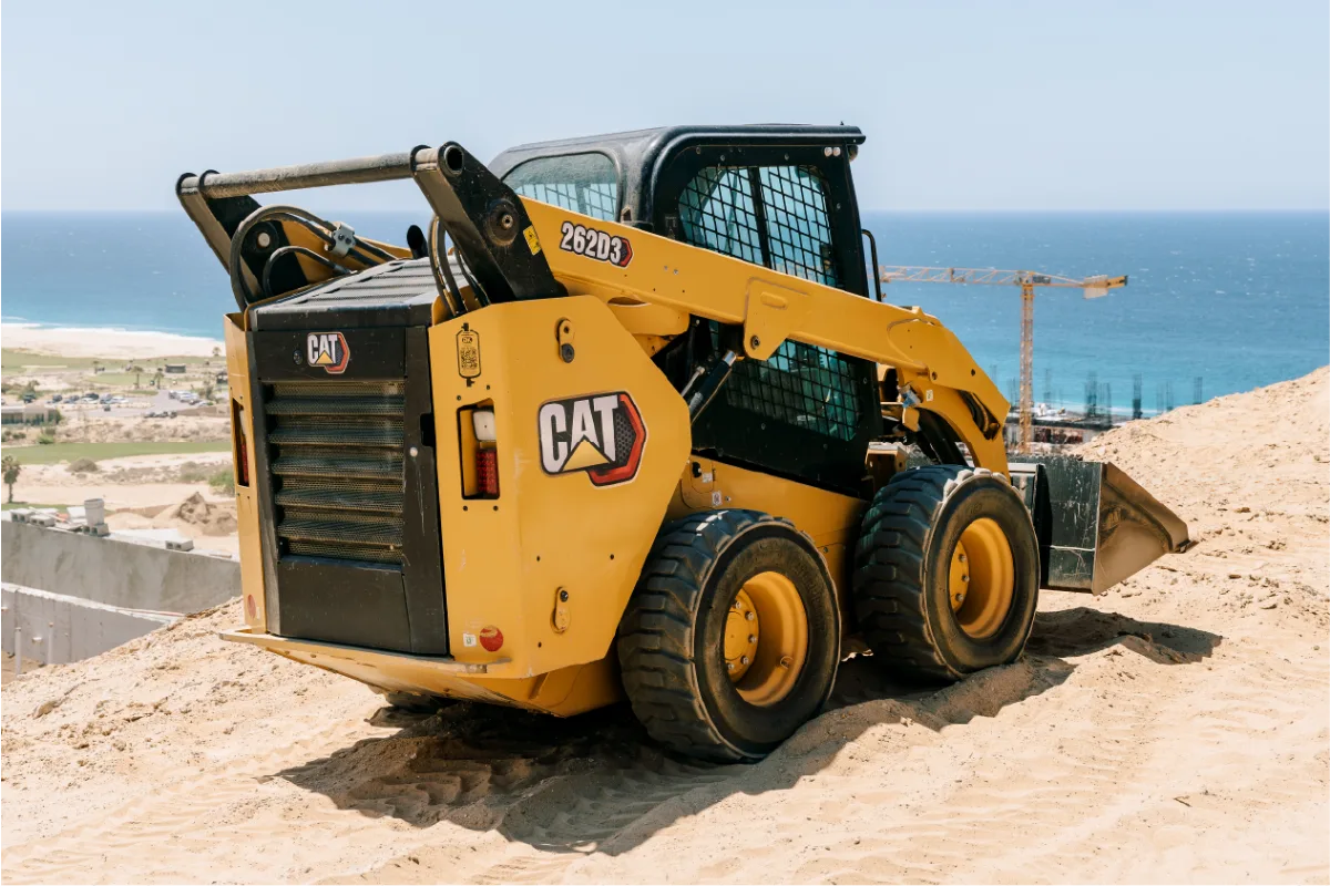 Rear three quarter view of the CAT 262D3 skid steer on a sand mound, showing its engine grille, 262D3 decal and rubber tires, with the ocean and a construction crane behind