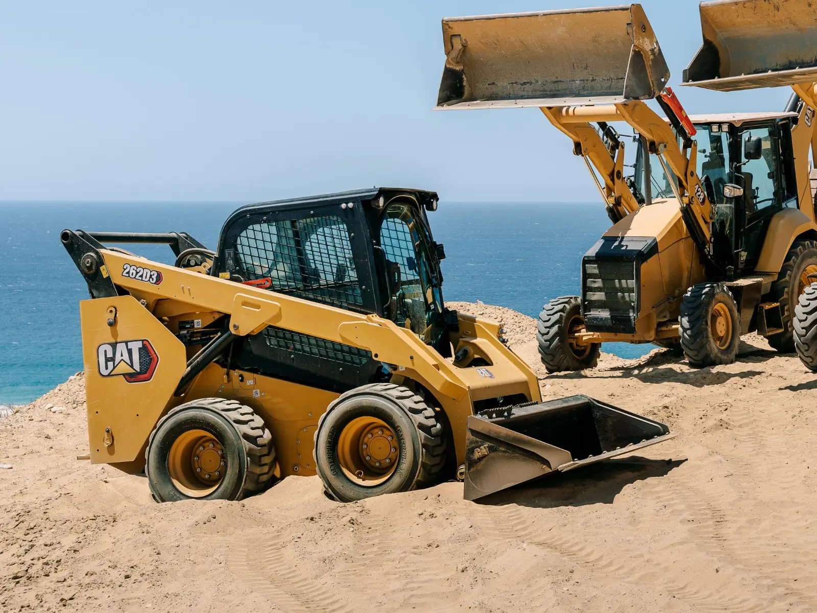 Yellow CAT 262D3 skid steer with its bucket resting in the sand on a coastal bluff, and two CAT backhoe loaders behind it lifting their front buckets against a blue sky
