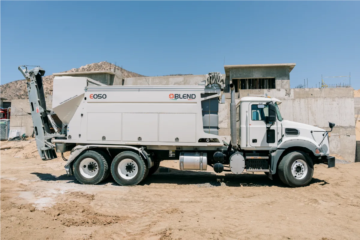 White Mack truck carrying the Blend E050 mixer body, photographed from the side with the discharge conveyor folded at the back, in front of a concrete house frame