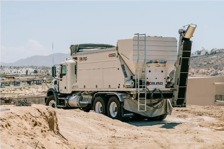 The Blend E050 on its Mack truck from behind and to one side, with the access ladder and the discharge conveyor raised above the hopper, houses and hills in the distance