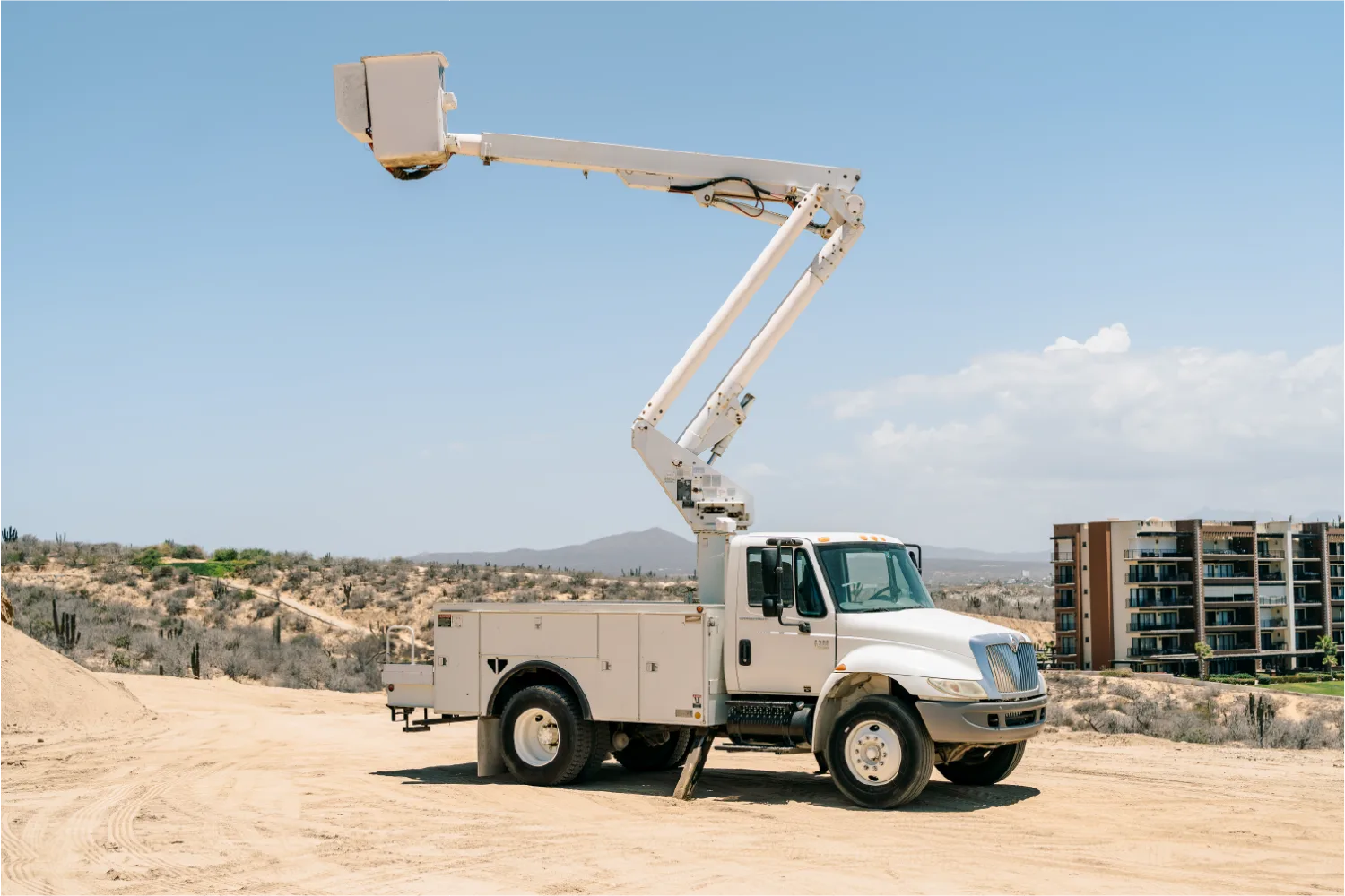 White International 4300 work truck with its boom raised and the personnel basket high above a sandy lot, desert hills and a condominium building behind