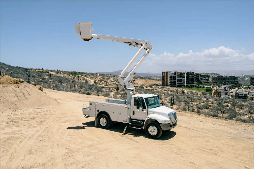 International 4300 basket lift truck on a graded lot with its folding boom stretched out above the cab and a stabilizer leg lowered, homes on the hillside beyond
