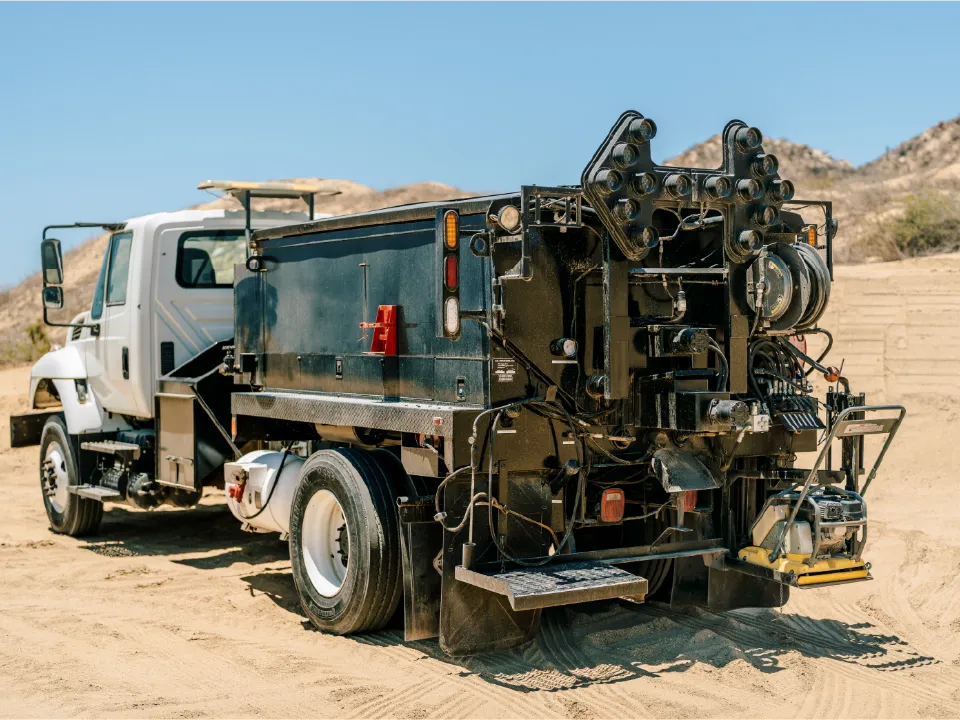 International work truck carrying a black road patching unit, parked on sand below desert hills