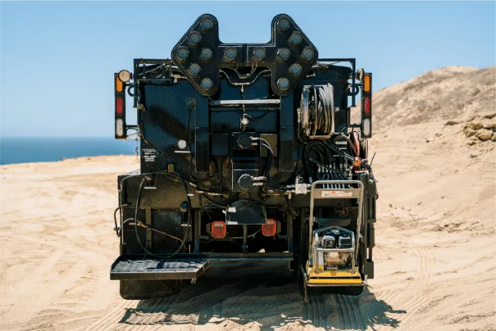 Rear of the patcher seen straight on, with the arrow board lights on top, hydraulic lines and a hose reel, the sea showing past a sandy slope