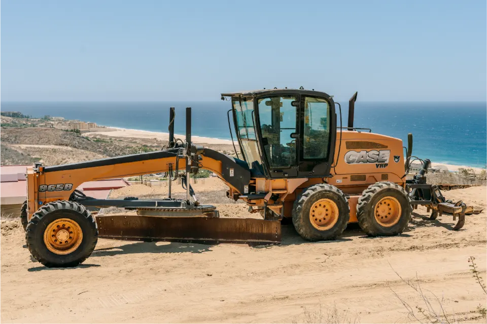 Blade lowered, a CASE VHP 865B grader waits high on a sandy bluff, a long curve of beach and blue water below