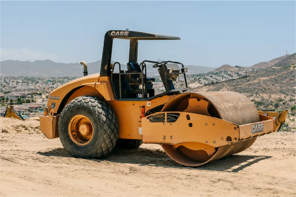 Orange CASE roller compactor parked on a sandy lot, a hillside covered in houses and distant mountains behind