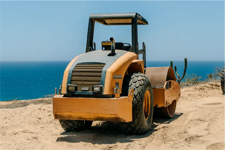 Front view of an orange CASE roller compactor with a single drum, set on a sandy hilltop with a deep blue ocean filling the background