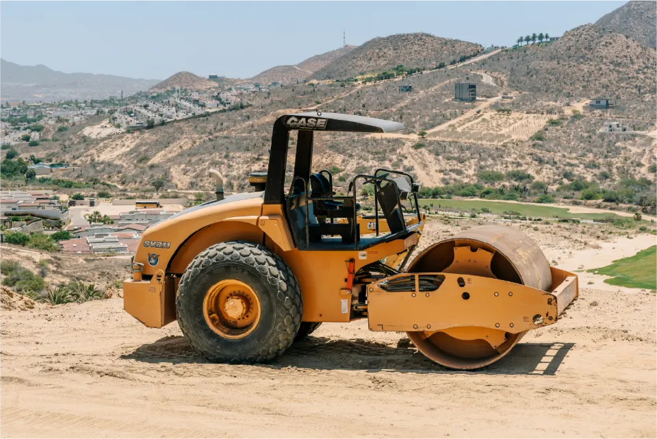Side view of an orange CASE single drum roller on a dirt pad, with homes, a green fairway and rugged hills in the valley behind