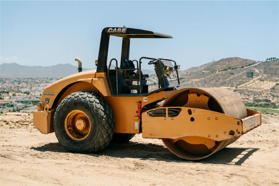 Angled view of an orange CASE roller compactor with a canopy over the seat, standing on a dirt pad above homes and hills