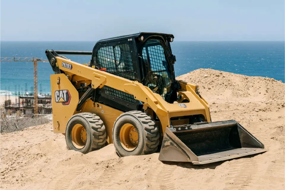 Bucket empty, the CAT 262D3 sits on a sandy ridge with the operator seated inside and blue water behind