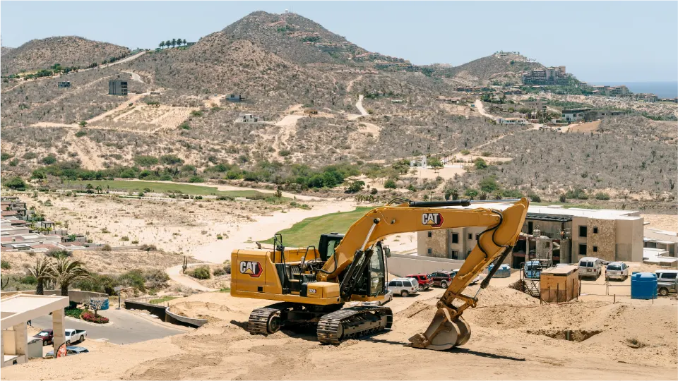 Wide view of a CAT 320GC excavator on a building lot, with desert mountains, a green fairway and the parking area of an apartment complex behind it