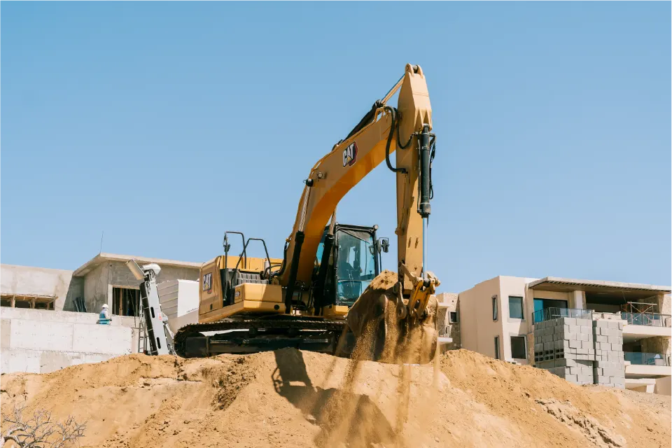 Head on view of a CAT 320GC excavator on top of a mound, sand falling from its bucket, unfinished apartment buildings on both sides