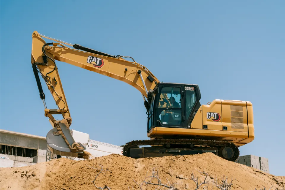 CAT 320GC excavator, operator visible in the glass cab, reaching down the far side of a sand mound with a Blend mixer truck parked behind