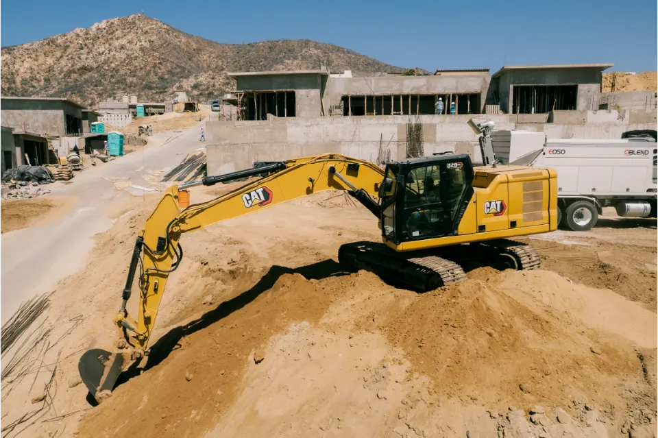 CAT 320GC excavator on a sand pile stretching its bucket downhill toward a paved street, house frames and a mountain behind