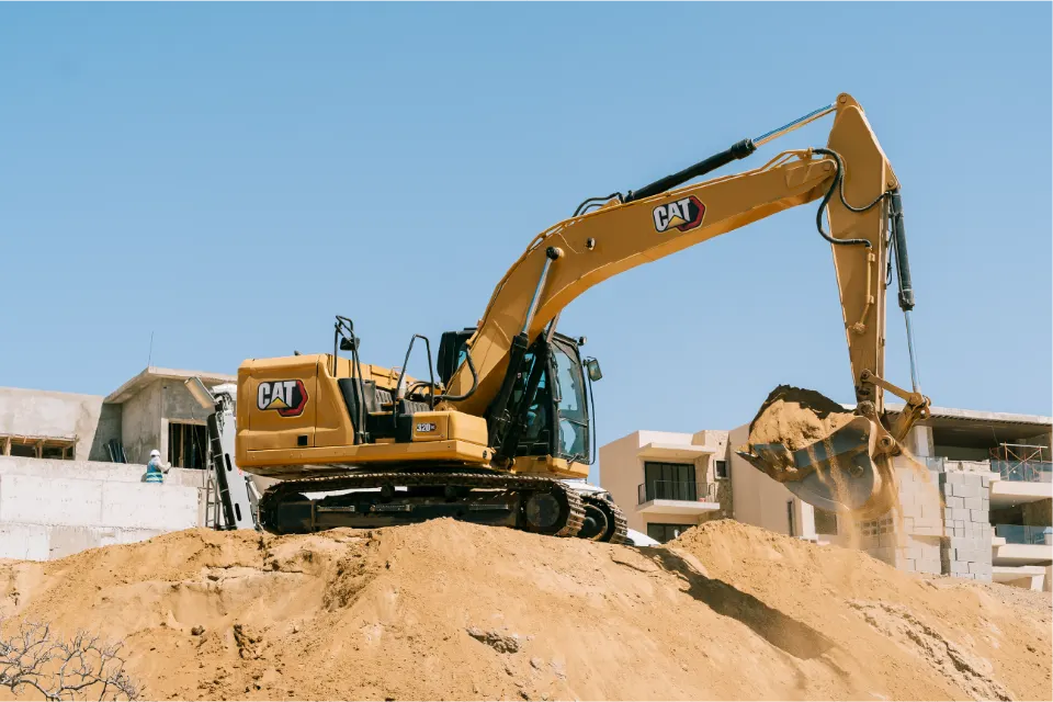 CAT 320GC excavator balanced on a sand pile, sand spilling out of its lifted bucket, with concrete apartment buildings under construction behind