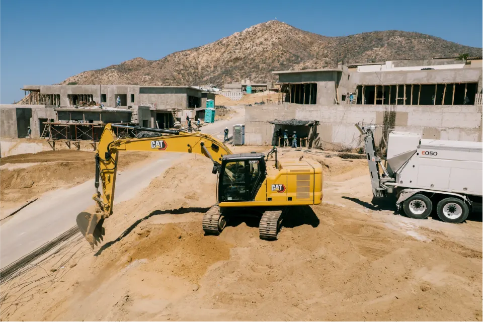 CAT 320GC excavator digging on a terraced hillside lot, concrete house frames and a Blend mixer truck beyond, a rocky peak above
