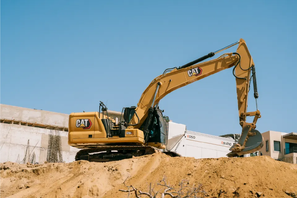 CAT 320GC excavator cresting a sand mound, bucket lowered, a mixer truck and building frames behind