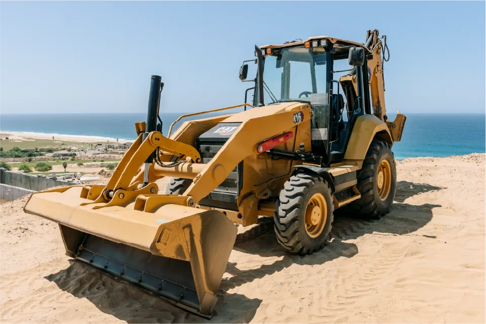 Front view of a CAT 416 backhoe loader, its loader bucket resting on the sand, with a golf course, buildings and the coast behind