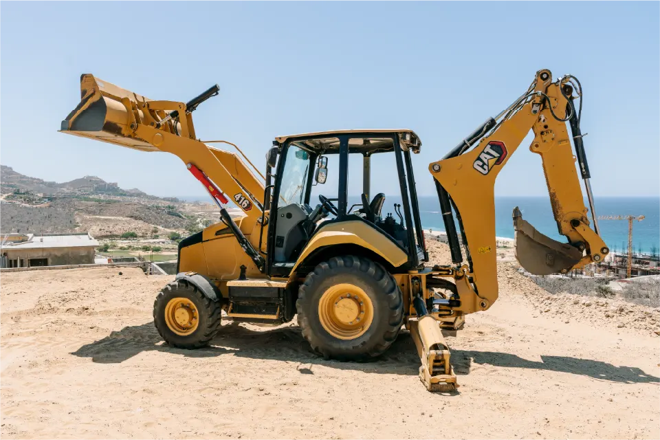 Side view of a CAT 416 backhoe loader standing on a sandy rise, both the loader bucket and the backhoe arm raised, with the coastline behind