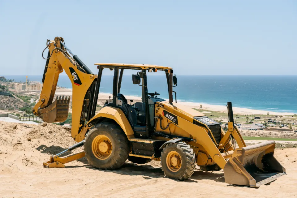 CAT 416F2 backhoe loader with its stabilizers down on a sandy bluff, the beach, a golf course and the ocean spread out below