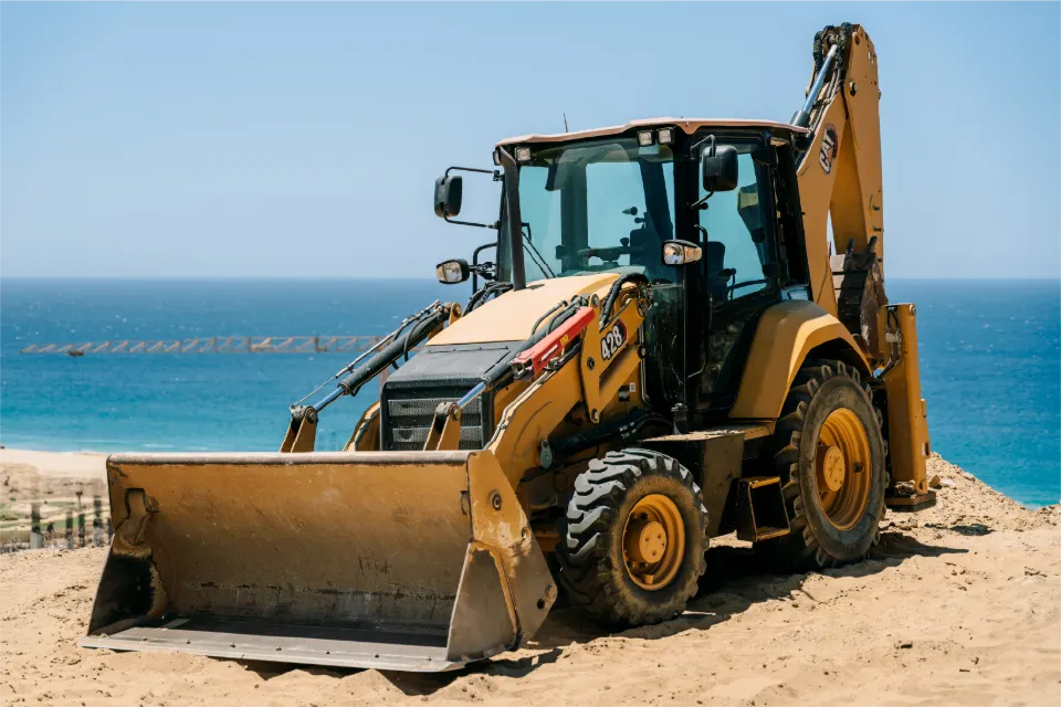 Front view of a CAT 428 backhoe loader, loader bucket set down, at a sandy edge with deep blue sea behind