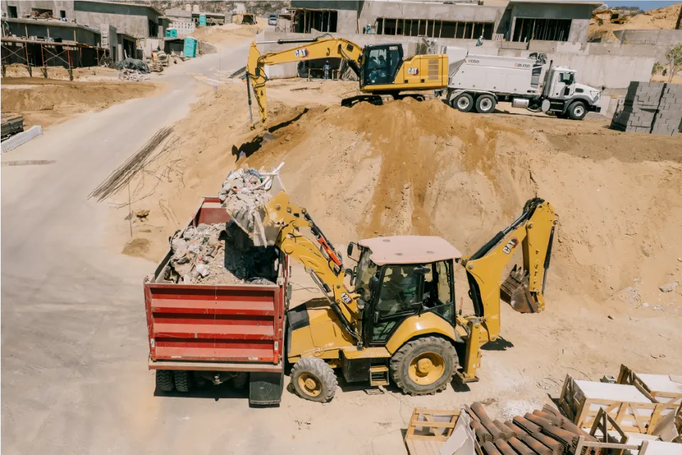 A CAT backhoe loader drops rubble into the red body of a dump truck while a CAT excavator and a mixer truck work farther up the hill among new houses
