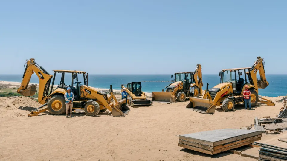 Four CAT backhoe loaders spread across a beach work area, with crew members in safety vests standing beside them and the sea behind