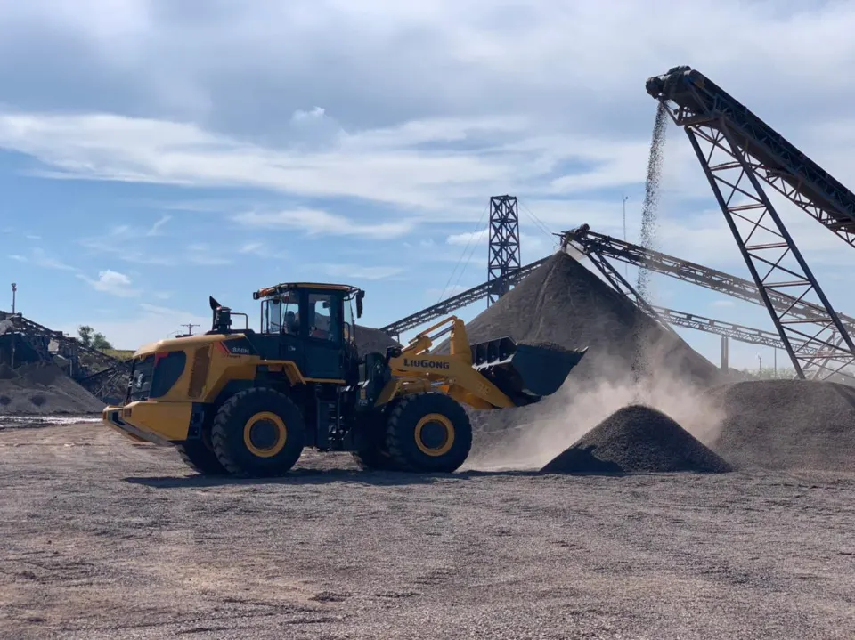 A yellow LiuGong 856H works in front of conveyor belts that drop crushed stone onto tall stockpiles, dust hanging in the air