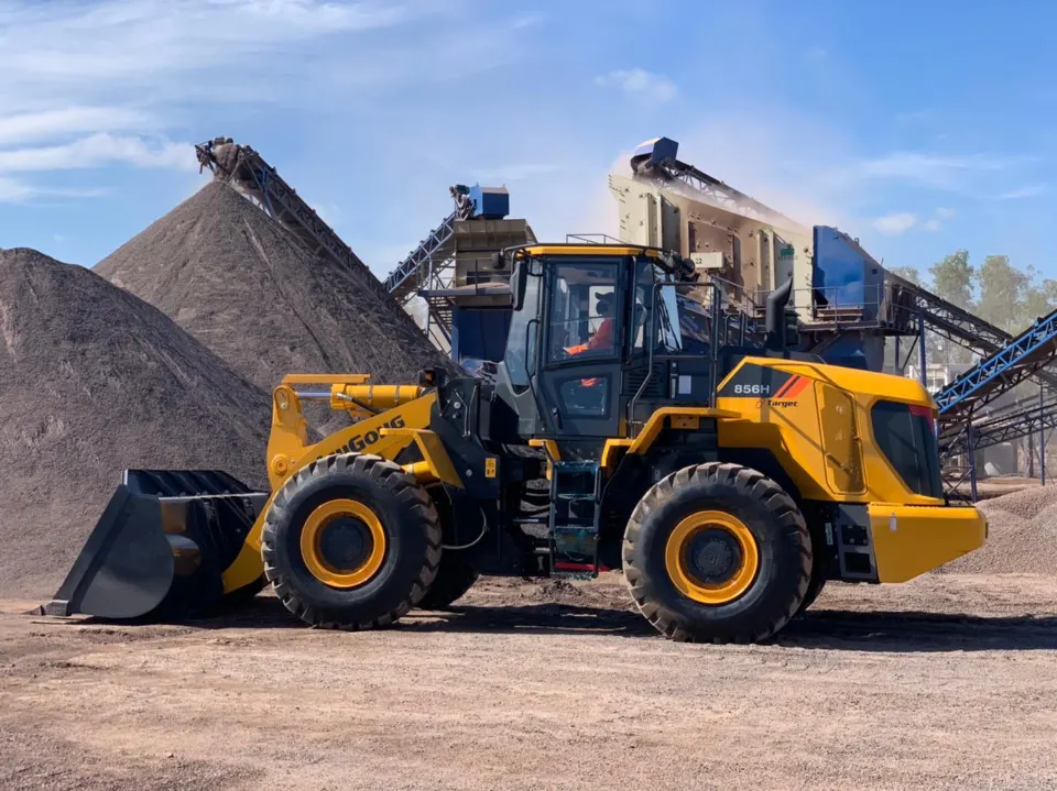 Side view of a LiuGong 856H, operator aboard, parked before tall gravel piles and a crushing plant