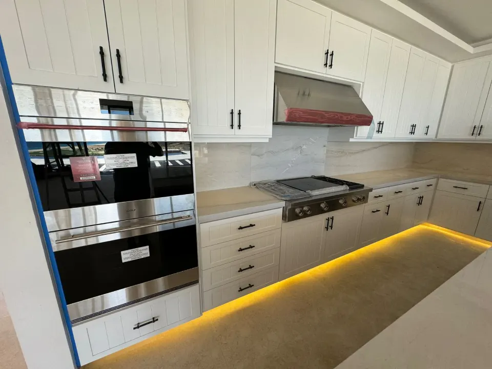 New kitchen with white shaker cabinets, a stainless range hood, a gas cooktop, wall ovens and warm strip lighting along the base cabinets