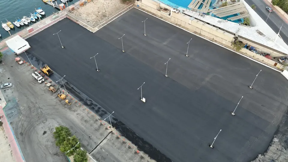 Seen from the air, a broad freshly paved lot with light poles beside a marina with small boats and a waterfront street
