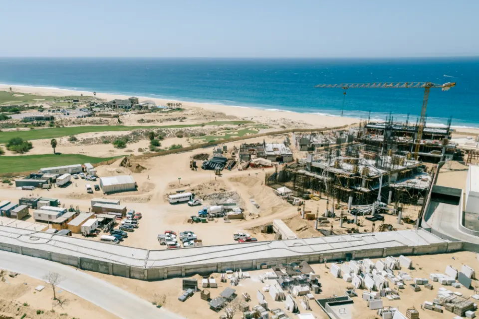 Aerial view over a large beachfront build: a tower crane, rows of concrete frames, parked trucks and material yards, with the ocean along the top of the frame