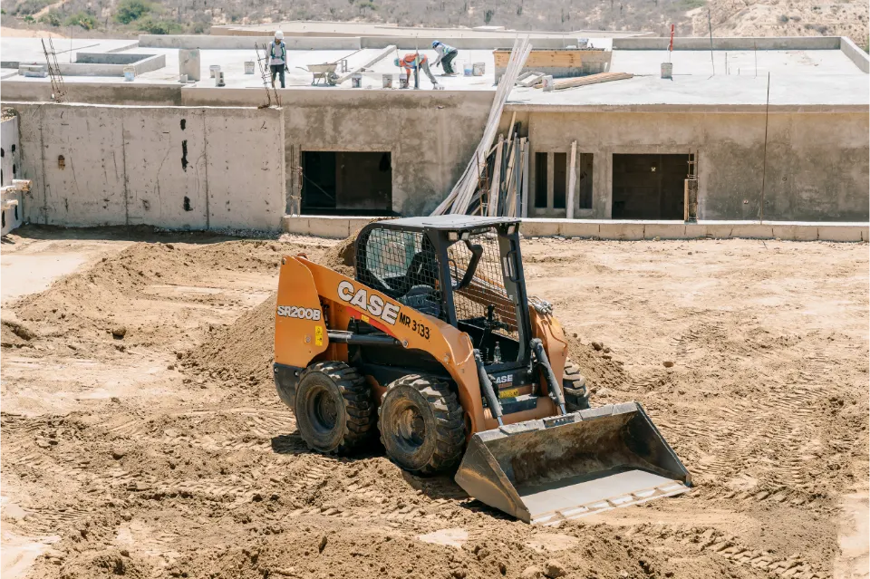 CASE SR200B skid steer with a loaded bucket before a concrete building shell where crew members work on the roof