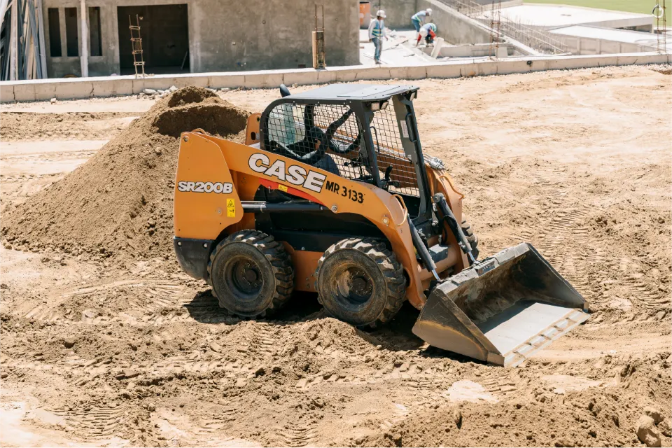 Orange CASE SR200B skid steer pushing a bucket of soil beside a dirt pile, with workers on a concrete structure behind