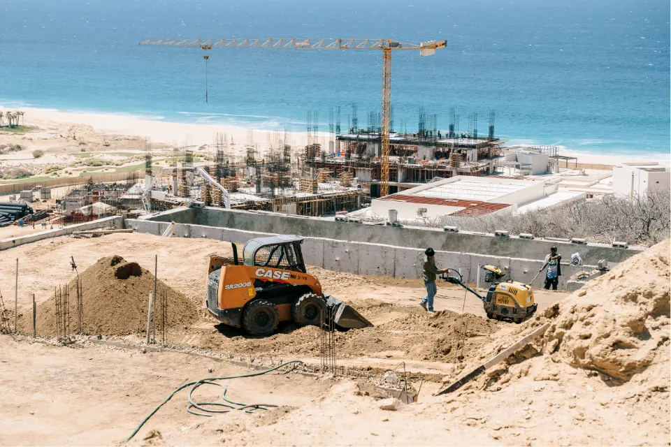 Orange CASE skid steer moving fill while two workers run a plate compactor, with a tower crane, a building frame and a white sand beach behind