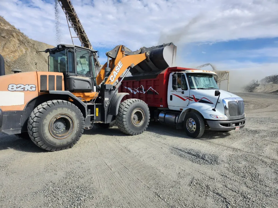 CASE 821G wheel loader tipping gravel into a red and white International dump truck on a gravel yard, a conveyor and a rocky slope behind