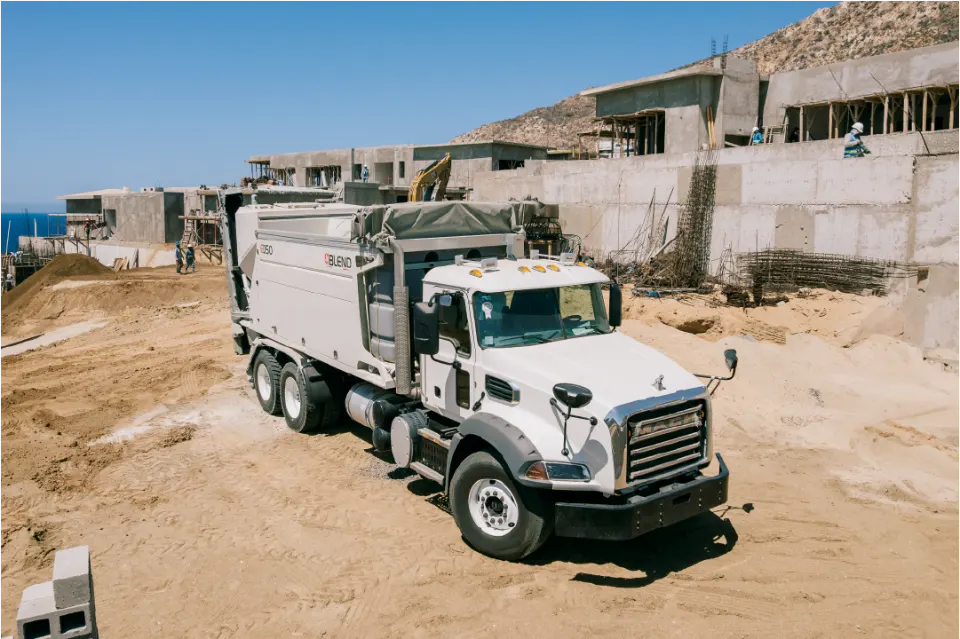 Angled front view of the white Mack with its Blend E050 mixer, parked on sand with concrete blocks and terraced house frames behind