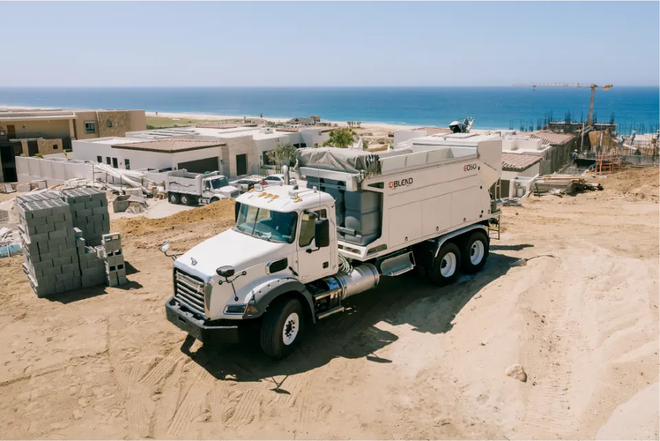 Elevated view of the Blend E050 mixer on its white Mack truck at a residential build, with stacked concrete blocks, unfinished houses and the sea behind