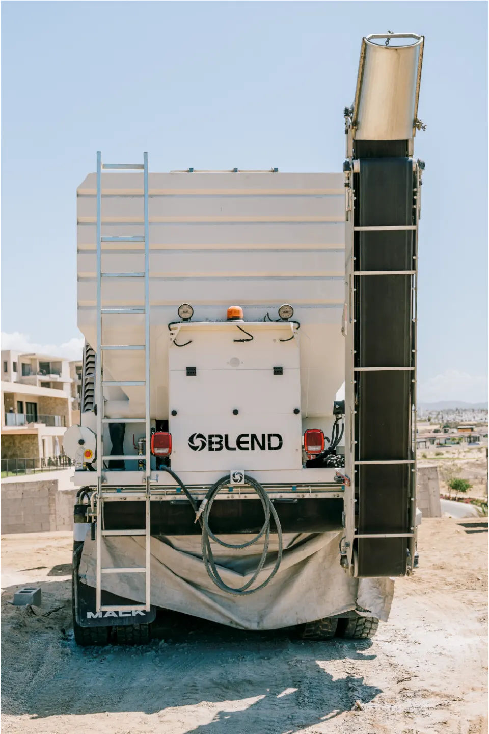 Rear view of the Blend E050 unit showing its ladder, control panel and tall discharge conveyor standing upright, apartment buildings behind