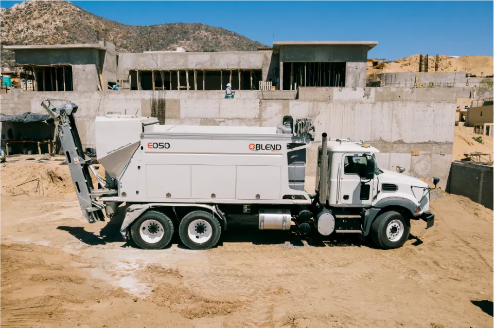 Blend E050 mixer on a Mack truck, conveyor raised, parked on graded sand in front of concrete retaining walls and house frames