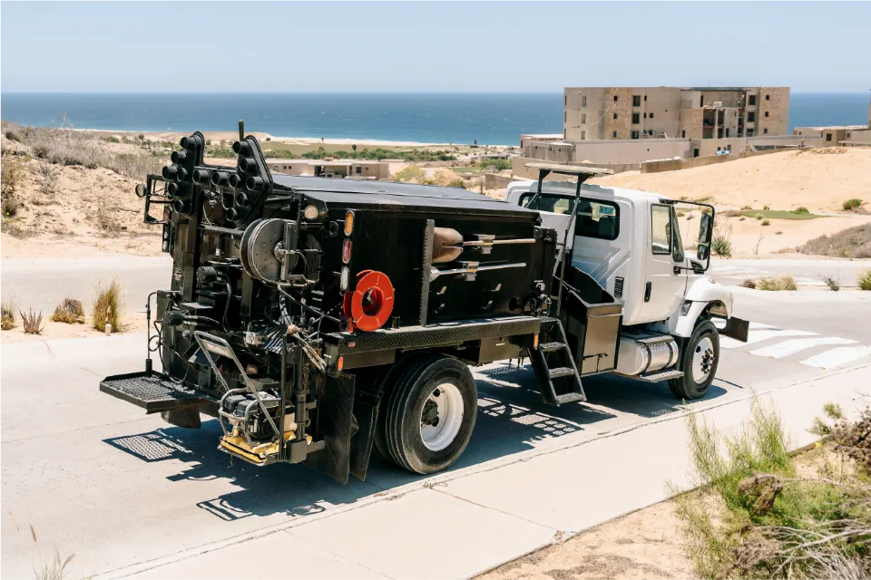 From the rear corner, the white International truck and its Pro Patch 4000 rig on a curving paved street, an unfinished building and the ocean beyond